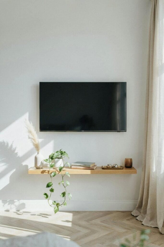 A white minimalist TV wall with a wall-mounted screen, slim floating shelf, and linen curtains diffusing morning light